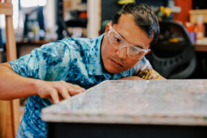 Navigating Home fellow Justin Cruz, who is assigned to the G3 Makerspace & Innovation Hub, checks the surface of the desk prototype that he created. The G3 Makerspace is collecting recyclable plastics to support a new project aimed at refurbishing school desks.