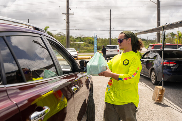 G3 Conservation Corps member handing bagged goods to a driver through a car window at a community giveaway.